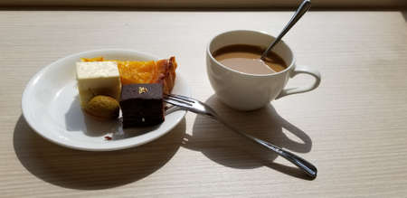 Close up image of small pieces of a peach pie, brownie, cookie, and a cheesecake on a small porcelain plate. A shiny fork is next to plate and all are served with a cup of milk coffee in a mug.の写真素材