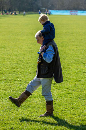 Oxford ,UK 03/19/2011: An elderly grandfather is carrying his grandson on his shoulders as they both stroll through the park in a sunny afternoon. They both wear boots and casual winter clothes.のeditorial素材