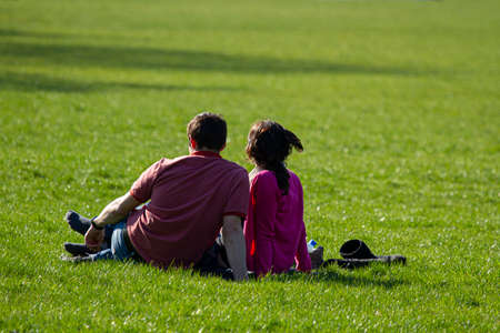 A young couple is having some leisure time as they sit on the grass and relax in a park on a sunny afternoon. Man wears a t-shirt and jeans, woman wears pink blouse and had her boots off at the side.の写真素材