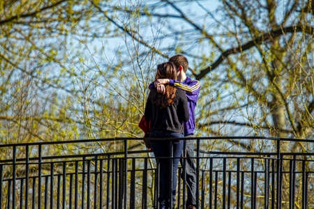 Oxford, UK 03/19/2011: A young couple is standing on a bridge at a park and cuddling each other. The boy wearing tracksuit has his arms around her shoulders as he is about to kiss her.のeditorial素材