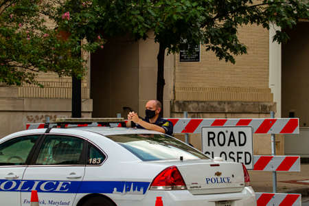Frederick, MD, USA 08/14/2020: A young caucasian male police officer is wearing a face mask while on duty. He is standing by his police car in front of a road block sign.のeditorial素材