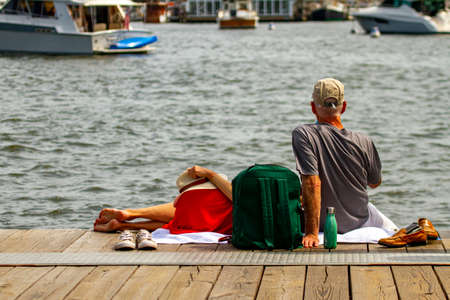 A caucasian couple is enjoying the nice weather on Annapolis. Both have taken off their shoes. The man sits with legs hanging off the wooden pier and the woman is lying on his lap.の写真素材