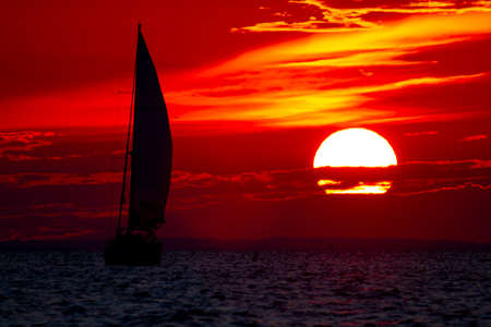 A spectacular sunset over Chesapeake Bay Maryland as captured from the east shore. Image features the large sun going down among clouds on a red sky and the silhouette of a sailboat with full sails.の写真素材