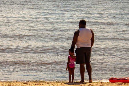 Rock Hall, MD, USA 08/30/2020: An African American man with white undershirt is standing on the beach next to her little daughter who is wearing a swimsuit. They look at the calm Chesapeake bay.のeditorial素材