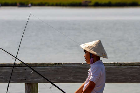 Eastern Neck Island, MD, USA  09/06/2020: An Asian boy wearing a traditional Chinese straw bamboo coolie cone hat is holding a fishing rod on a bridge over an estuary. He is fishing on a sunny  day.のeditorial素材