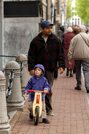 Amsterdam, Netherlands 05/15/2010: A middle eastern or Indian father is looking after his son who is riding a bike outside on the sidewalk at a busy urban location.のeditorial素材