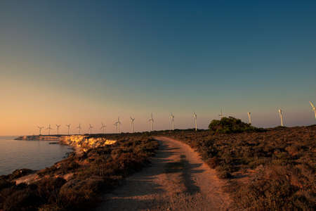 A landscape image taken on the western cape  of Bozcaada, Turkey. Image shows the scenic wind farm, a collection of wind turbines, at sunset. Also seen are cliffs, a dirt road and maquis shrubland.の写真素材