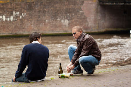 Amsterdam, Netherlands 05/15/2010: Two young men are sitting on concrete floor and having a store bought cold snack with a bottle of wine by the river on a weekend day.のeditorial素材