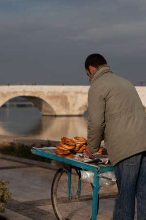 A young man is selling simit (Turkish sesame bagel) on a wooden food cart in the street. Historic...