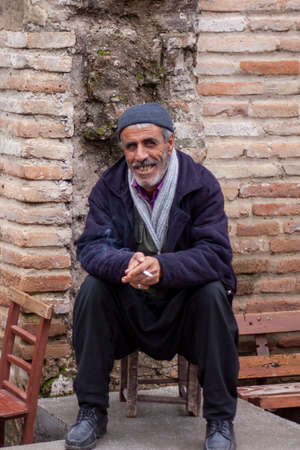 Adana, Turkey 12/26/2009: portrait of an elderly Turkish man wearing salwar and a jacket and sitting on a wooden chair while smiling and smoking a cigarette. Image was taken at the old town of Adanaのeditorial素材
