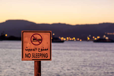 Aqaba, Jordan 03/31/2010: Close up image of a "No Sleeping" sign on a wooden post that is aimed to prevent people sleeping on the beach. Red Sea, mountains, city lights of Eilat are seen in backgroundのeditorial素材