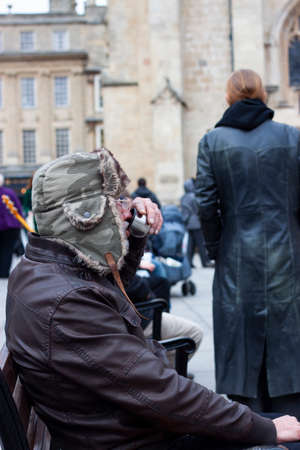 Bath, UK 03/06/2010:  A funny old man who is holding a metal cup of hot drink and a cigarette while sitting on a bench is sticking out his tongue for mockery. He is wearing wooly trapper hat.のeditorial素材