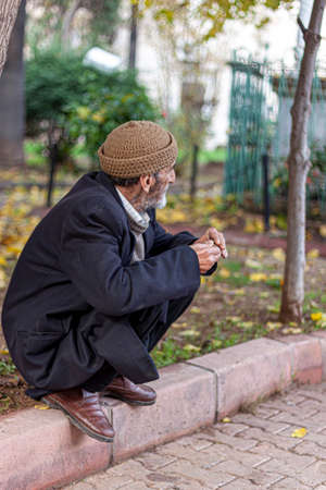 Adana, Turkey, 12/31/2009: An elderly man wearing traditional hat, winter coat, and leather shoes is squatting on the edge of a sidewalk. He is looking away while rubbing his hands together.のeditorial素材