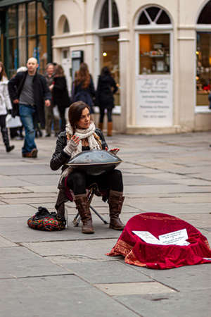 Bath, UK, 03/06/2010: A young woman musician is sitting on a foldable stool in a crowded street and playing hang instrument. This street performer is also selling her original music recordings.のeditorial素材
