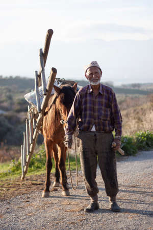 Manisa, Turkey 12/31/2009: Elderly villager wearing old clothes and a hat is pulling a horse that carries a wooden ladder and some bags. He looks tired as he walks through the unpaved mountain path.のeditorial素材