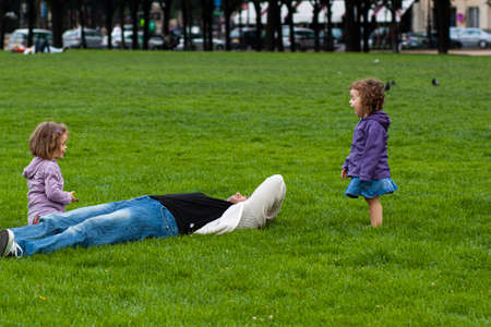 Paris/France 06/13/2010: A father is lying on grass in an outdoor park. He is trying to rest or sleep but his two little daughters do not leave him alone. They are waking him up to play.