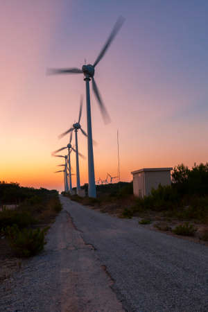 Close up image of the  wind turbines at Bozcaada wind farm. Long exposure shot captures the turbines as they spin. A dirt road and  a transformer building are seen. Maquis shrubs are in the backgroundの写真素材