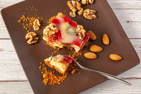 A slice of fruit cheesecake served on a brown porcelain plate with walnuts and almonds. The cake has raspberry and kiwi fruits which is finished with custard icing. A metal fork separates a bite size.の写真素材