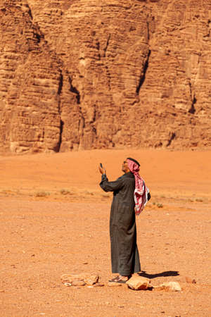 Wadi Rum, Jordan 04/01/2010: An Arabic man wearing traditional keffiyeh, agal, sunglasses and Jubba thobe (long dress) is lifting up his mobile phone in air to search for reception in desertのeditorial素材
