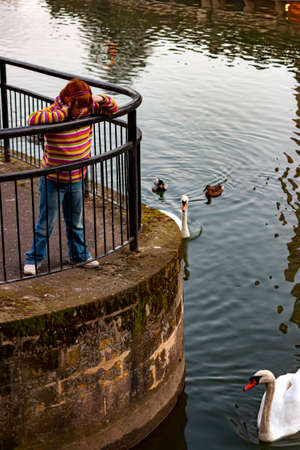 Bath, UK 03/06/2010: A school aged caucasian girl with red hair wearing colorful sweater and jeans is leaning on metal railings by Avon river and looking down at the swans swimming.  by the mossy wallのeditorial素材