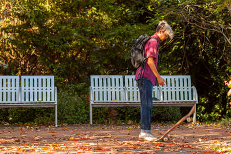 Frederick, MD, USA 10/14/2020: A teen boy with stylish blond hair wearing jeans and sneakers is stepping on the tail of his skateboard to pop it in the air. He is on a road covered with autumn leavesのeditorial素材
