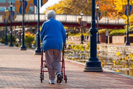 An elderly caucasian woman wearing blue sweater, track pants and sneakers is slowly walking in parks...