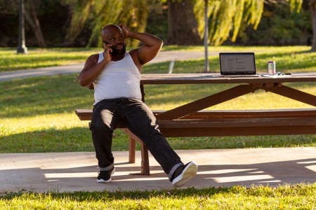 Frederick, MD, USA 10/14/2020: An African American young man wearing a white undershirt is sitting on a picnic table at Baker Park and is talking on the phone while stretching legs and scratching headのeditorial素材