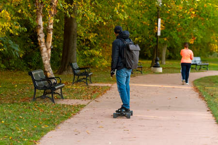 Young man wearing hoodie, backpack and a hat is skateboarding on a concrete path in Baker Park....