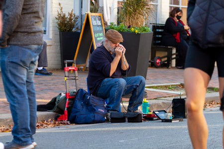 Alexandria, VA, USA 11-28-2020: A white bearded elderly street musician wearing boots, jeans and flat hat sitting on stool by the sidewalk is playing harmonica attached to a speaker for donations.のeditorial素材