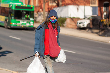 Frederick, Maryland, USA 11-22-2020: A long haired caucasian homeless man is walking in the street in downtown Frederick wearing dirty old winter clothes. He carries his belongings in white trash bagsのeditorial素材