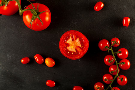 A flat lay image featuring a medley of tomatoes on black wooden background. Some Roma tomatoes and cherry tomatoes are on vine, one of them is sliced in half. Healthy and fresh eating conceptの写真素材