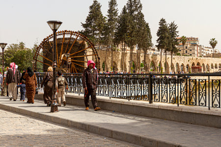 Hama, Syria 04-02-2010: Cityscape of the historic city of Hama featuring a bridge over Orontes river where people in ethnic clothes are walking. In the background there is a noria and aqueduct.のeditorial素材