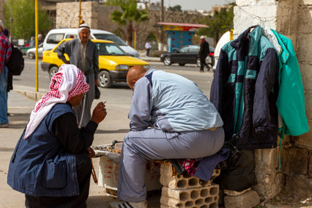 Hama, Syria 04-02-2010: Two Syrian men are talking in the street. One is sitting on a brick trying to sell beads and small items on his small display. The other wearing keffiyeh is smoking cigarette.のeditorial素材