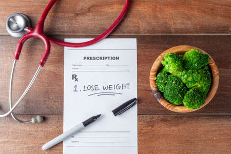 Close up flat lay image of a doctor's office desk with a bowl of broccoli,  stethoscope, pen and a prescription that has one item "Lose weight" prescribed. A concept image for obesity, diet, exerciseの写真素材
