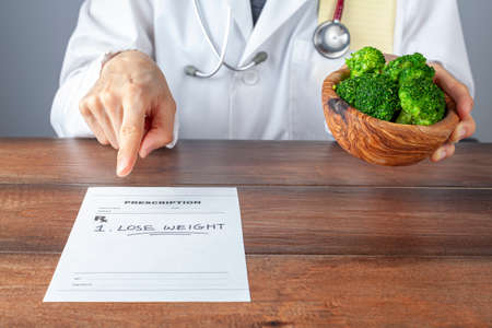 A caucasian female physician in white coat is pointing a prescription saying lose weight. She also offers a bowl of broccoli. Obesity, overweight, reduced calorie diet, lifestyle modification conceptsの写真素材