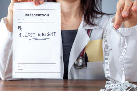 A female physician is holding a prescription that says lose weight in one hand and a tape measure...