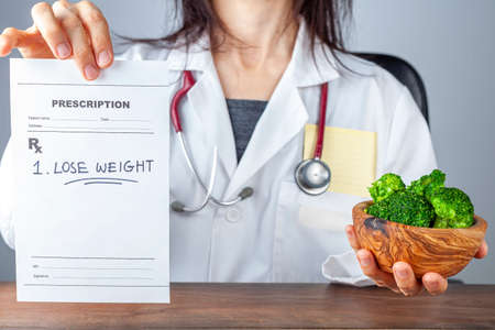 A caucasian female physician in white coat  is holding a prescription saying lose weight. She also offers a bowl of broccoli. Obesity, overweight, reduced calorie diet, lifestyle modification conceptsの写真素材