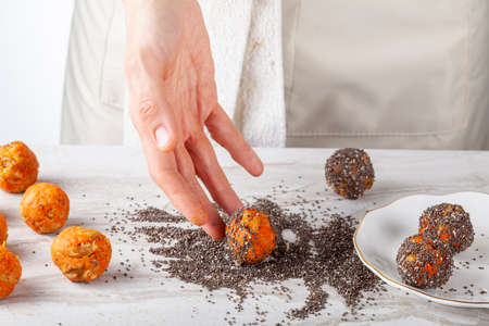 A woman chef is preparing carrot cake bliss bites in the kitchen. She dips the carrot balls into a pile of chia seeds and roll them to coat their surface. Then she moves them to plate to serve.の写真素材