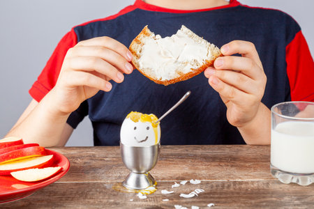 Breakfast table with simple balanced nutritious ingredients. A soft boiled egg with face painted on, a slice of toasted bread with cream cheese spread, apple, a glass of milk and a boy eating thereの写真素材