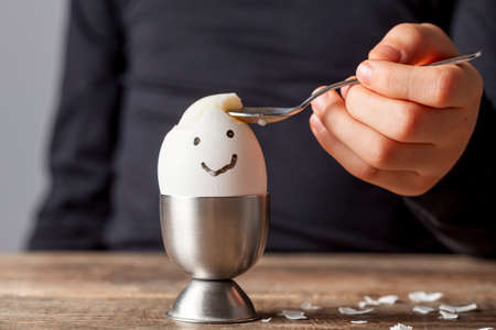A small child is eating soft boiled humpty dumpty egg placed in egg cup on a wooden breakfast table. She uses spoon to eat the egg from its shell. She has drawn a smiling face on the shell.の写真素材