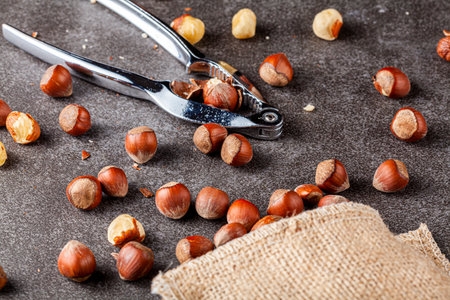 closeup selective focus image of hazelnuts in shells scattered from a vintage burlap sack onto dark background. Some are shelled and some are cracked. A metal nut cracker is there to crack open nuts.の写真素材