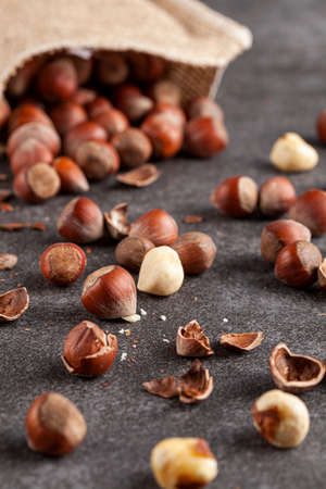 closeup selective focus image of hazelnuts in shells scattered from a vintage burlap sack onto dark background. Some are shelled and some are cracked. A metal nut cracker is there to crack open nuts.の写真素材
