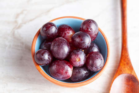 A simple image of a porcelain bowl or red grapes  and a wooden spoon on marble countertop. A concept...