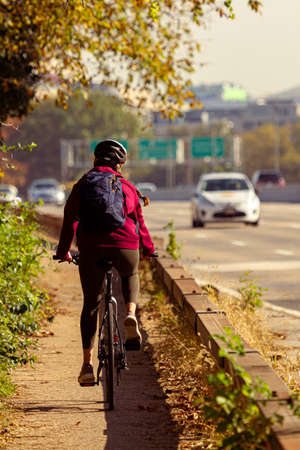 A young woman wearing red hoodie, helmet, tights and sneakers is cycling on a narrow bike path nearby a large highway in Washington DC. She is commuting with the bicycle and her backpack is with her.の写真素材