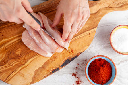 Close up image of a woman chef slicing chicken breast to male stripes and nuggets. She cuts the meat on wooden chopping board using sharp knife. Cayenne red pepper and salt are on side for seasoningの写真素材