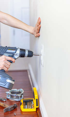 A woman is using a cordless electrical drill with screwdriver bit attached to remove the screw holding the cover plate of an electrical outlet on the wall during a DIY project. Toolkit on wooden floorの写真素材