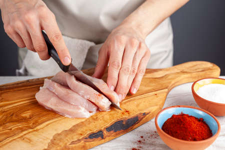 Close up image of a woman chef slicing chicken breast to male stripes and nuggets. She cuts the meat on wooden chopping board using sharp knife. Cayenne red pepper and salt are on side for seasoningの写真素材