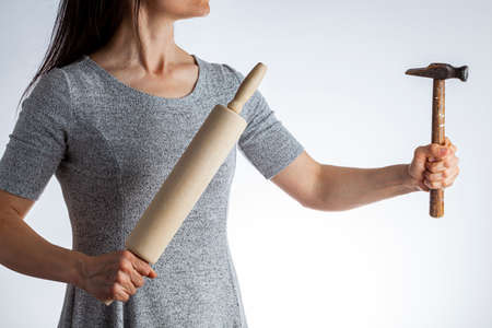 A young caucasian woman wearing short sleeve dress is holding a hammer and rolling pin in her hands....