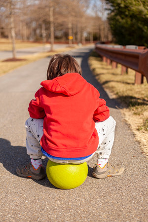 A little child wearing red hoodie and sneakers is resting on a ball after getting tired at a game. The person is alone in a hiking path at a rural location.の写真素材