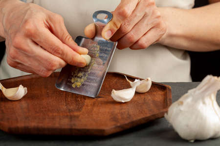Hands of a caucasian woman grating garlic cloves using shovel shaped metal mini grater on a wooden plate. This small item can also be used as ginger grater. A convenient durable tool.の写真素材